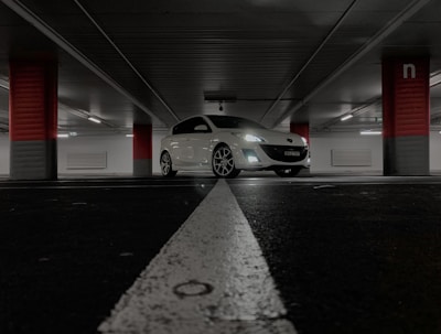 A white car is parked in an underground parking garage. The environment is dimly lit, with fluorescent lights creating a subdued atmosphere. Red and gray pillars support the ceiling, and white markings divide the parking spaces.