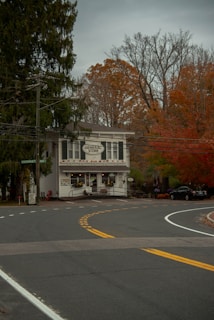 A quaint general store is situated at the curve of a road, surrounded by autumn trees with orange and red leaves. The storefront features a wide entrance with decorative elements and signs. The scene is calm, with overcast skies contributing to a rustic, cozy atmosphere.