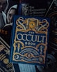 A collection of books with themes related to the occult, witchcraft, and alchemy. The focal point is a book titled 'The Occult Book' by John Michael Greer, featuring ornate gold lettering and symbolic decorations on a dark blue cover. Surrounding the main book are other titles concerning witchcraft, demonology, and magic spells. The overall design is dark and mystical.