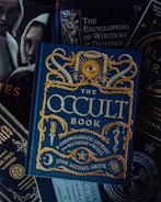 A collection of books with themes related to the occult, witchcraft, and alchemy. The focal point is a book titled 'The Occult Book' by John Michael Greer, featuring ornate gold lettering and symbolic decorations on a dark blue cover. Surrounding the main book are other titles concerning witchcraft, demonology, and magic spells. The overall design is dark and mystical.