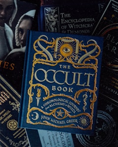A collection of books with themes related to the occult, witchcraft, and alchemy. The focal point is a book titled 'The Occult Book' by John Michael Greer, featuring ornate gold lettering and symbolic decorations on a dark blue cover. Surrounding the main book are other titles concerning witchcraft, demonology, and magic spells. The overall design is dark and mystical.
