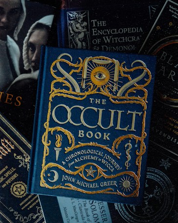A collection of books with themes related to the occult, witchcraft, and alchemy. The focal point is a book titled 'The Occult Book' by John Michael Greer, featuring ornate gold lettering and symbolic decorations on a dark blue cover. Surrounding the main book are other titles concerning witchcraft, demonology, and magic spells. The overall design is dark and mystical.