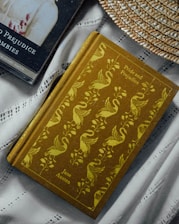 Two books are placed on a fabric surface. One book, 'Pride and Prejudice' by Jane Austen, has an ornate cover featuring a pattern of swans and foliage in yellow on a brown background. The other book is 'Pride and Prejudice and Zombies' with a dark cover showing part of a character. A woven straw hat is partially visible, adding a rustic element to the composition.