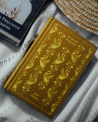 Two books are placed on a fabric surface. One book, 'Pride and Prejudice' by Jane Austen, has an ornate cover featuring a pattern of swans and foliage in yellow on a brown background. The other book is 'Pride and Prejudice and Zombies' with a dark cover showing part of a character. A woven straw hat is partially visible, adding a rustic element to the composition.