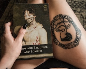 A hand with black nail polish is holding a book titled 'Pride and Prejudice and Zombies.' The book cover features a classical painting of a woman with a zombie twist, showing skeletal features and blood. Next to it, there is a thigh tattoo of an ornate frame with a woman in Victorian attire.