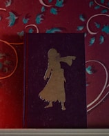 A book with a dark cover features a gold silhouette of a person wearing a long dress. The book is placed against a wall with a floral pattern in red, with blue and teal leaves and white swirls.