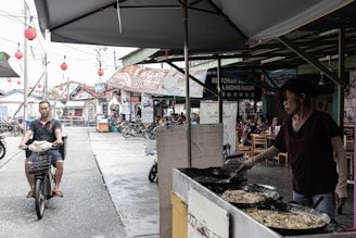 A bustling street scene with a food vendor cooking in large pans under an umbrella. A man rides a motorbike along the street lined with shops and red lanterns hanging above. Several bicycles are parked and people are seated at tables in the background, indicating a lively area.
