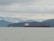 A large cargo ship travels across a calm body of water with a backdrop of forested hills and misty mountains under an overcast sky. A small lighthouse is visible on the shoreline to the left.
