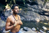 A man is meditating with his eyes closed near a tranquil stream. He is shirtless and wearing a beaded necklace and a colorful bracelet, standing amidst a natural, rocky environment with greenery.