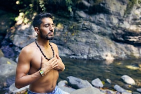 topless man wearing black beaded necklace and blue denim shorts standing on rocky shore during daytime