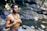 A man is meditating with his eyes closed near a tranquil stream. He is shirtless and wearing a beaded necklace and a colorful bracelet, standing amidst a natural, rocky environment with greenery.
