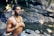 A man is meditating with his eyes closed near a tranquil stream. He is shirtless and wearing a beaded necklace and a colorful bracelet, standing amidst a natural, rocky environment with greenery.