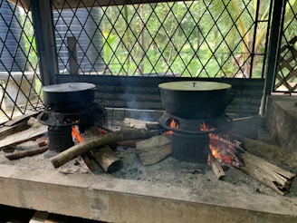 A rustic outdoor cooking setup features two large pots covered with lids, resting on metal stoves with wood logs used as fuel. The fires are burning brightly, emitting smoke under a canopy structure with a crisscross metal pattern. The background shows vegetation, including palm leaves and greenery.