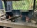 A rustic outdoor cooking setup features two large pots covered with lids, resting on metal stoves with wood logs used as fuel. The fires are burning brightly, emitting smoke under a canopy structure with a crisscross metal pattern. The background shows vegetation, including palm leaves and greenery.