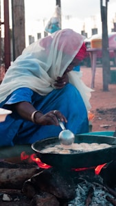 A person wearing vibrant traditional clothing and a headscarf is crouched down, cooking food over an open flame. They are using a ladle to stir or scoop something in a pan placed on a fire made of wood logs. The scene appears to be outdoors with a rustic setting.