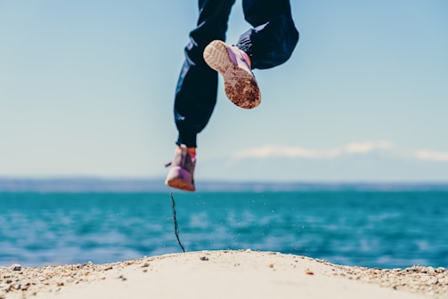 Close-up of a runner's feet pounding desert sand, kicking up dust under a bright blue sky.