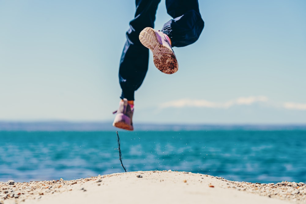 person in black pants and brown shoes jumping on brown rock near body of water during