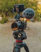 A professional camera is mounted on a sturdy tripod, equipped with a microphone and a monitor. The setup is situated outdoors with a blurred background of greenery, suggesting a natural setting. The camera and accessories have a sleek, high-tech appearance.