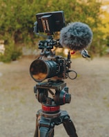 A professional camera is mounted on a sturdy tripod, equipped with a microphone and a monitor. The setup is situated outdoors with a blurred background of greenery, suggesting a natural setting. The camera and accessories have a sleek, high-tech appearance.