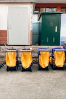 Four yellow cleaning carts with blue frames are aligned in front of a building with a white door and a green utility box. The background features a stone wall and some utility fixtures.