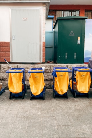 Four yellow cleaning carts with blue frames are aligned in front of a building with a white door and a green utility box. The background features a stone wall and some utility fixtures.