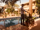 A man is standing near a swimming pool in a sunlit, lush garden setting. Surrounding the pool are potted plants and trees providing shade and greenery. The architecture of nearby buildings features wooden beams and a tiled roof, creating a warm, inviting atmosphere.