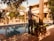 A man is standing near a swimming pool in a sunlit, lush garden setting. Surrounding the pool are potted plants and trees providing shade and greenery. The architecture of nearby buildings features wooden beams and a tiled roof, creating a warm, inviting atmosphere.