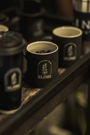 Multiple dark-colored mugs with the logo 'BLING' are placed on a wooden tray. One of the mugs contains coffee beans, and the setting appears to be dimly lit, giving a cozy and rustic feel.