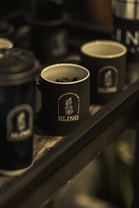 Multiple dark-colored mugs with the logo 'BLING' are placed on a wooden tray. One of the mugs contains coffee beans, and the setting appears to be dimly lit, giving a cozy and rustic feel.