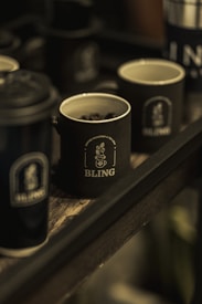 Multiple dark-colored mugs with the logo 'BLING' are placed on a wooden tray. One of the mugs contains coffee beans, and the setting appears to be dimly lit, giving a cozy and rustic feel.