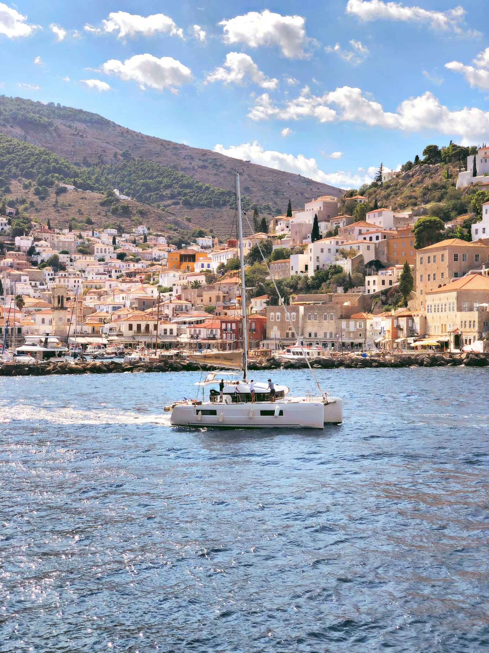 Stone mansions rising around Hydra's harbor as a sailing boat crosses the bay