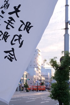 A Japanese street scene featuring a large white flag with black Japanese characters in the foreground. In the background, there are modern buildings, a tree, and a red train moving along the street under a partly cloudy sky.