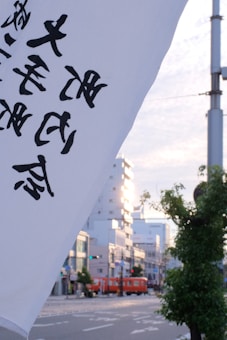 A Japanese street scene featuring a large white flag with black Japanese characters in the foreground. In the background, there are modern buildings, a tree, and a red train moving along the street under a partly cloudy sky.