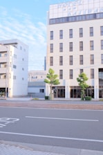 A tall building with a large hotel sign at the top is situated by the side of an empty street. The building's facade is light-colored, featuring numerous windows, and two small trees are planted on the pavement in front. The sky is clear with a few clouds, giving a calm and serene atmosphere.