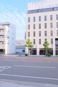 A tall building with a large hotel sign at the top is situated by the side of an empty street. The building's facade is light-colored, featuring numerous windows, and two small trees are planted on the pavement in front. The sky is clear with a few clouds, giving a calm and serene atmosphere.