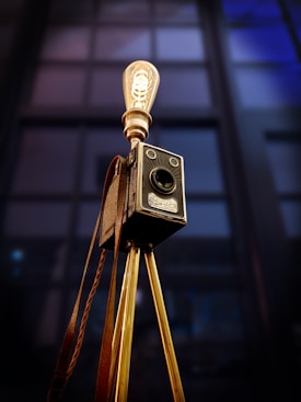 A vintage camera mounted on a tripod is repurposed into a lamp. The camera is detailed with leather accents and metal components, and the setup features a retro-style light bulb. The background appears to be tall windows, possibly suggesting an indoor or studio setting.