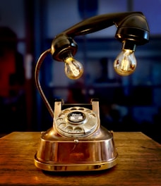 A vintage rotary dial telephone is creatively repurposed into a lamp with two exposed filament bulbs, one on each side of the receiver. The telephone is situated on a wooden surface with a dark, blurred background, giving a warm and nostalgic feel.
