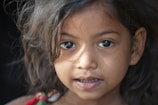 A close-up portrait of a young girl with dark, wavy hair. She gazes directly at the camera with large, expressive eyes. The lighting creates soft shadows on her face, highlighting her delicate features.