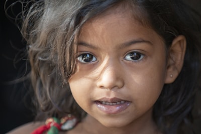 A close-up portrait of a young girl with dark, wavy hair. She gazes directly at the camera with large, expressive eyes. The lighting creates soft shadows on her face, highlighting her delicate features.
