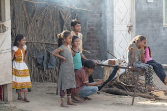 A group of children are gathered outside, engaging in what appears to be play or conversation. They are dressed in casual clothes, and their expressions suggest they are enjoying themselves. The background includes a rustic setting with elements like a wooden fence or structure, a metal door, and what seems to be a goat tethered nearby.