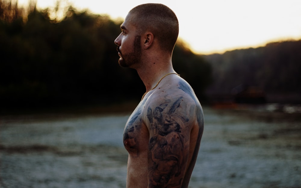 homme aux seins nus avec tatouage de corps debout près du plan d’eau pendant la journée