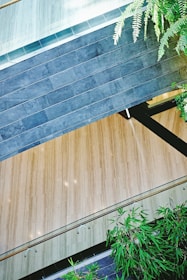A modern architectural space featuring a combination of wood and stone elements. Dark gray stonework contrasts with light wooden flooring, and there are transparent railings running along the edges. Lush green plants add a touch of nature to the environment, creating a harmonious blend of natural and architectural features.