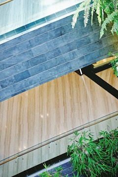 A modern architectural space featuring a combination of wood and stone elements. Dark gray stonework contrasts with light wooden flooring, and there are transparent railings running along the edges. Lush green plants add a touch of nature to the environment, creating a harmonious blend of natural and architectural features.