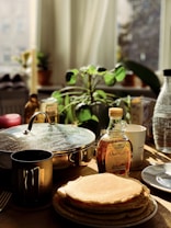 A cozy kitchen table setting with a stack of pancakes on a plate, alongside a bottle of maple syrup labeled 'Ahornsirup'. There's a metal travel mug, a covered pot with a shiny lid, and a plant with green leaves in the background. Soft sunlight filters through the window, casting warm, natural light.