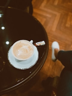 A cup of coffee sits on a white saucer placed on a glossy black table, accompanied by a small wrapper labeled 'Biscolata' Milano. The floor has a herringbone wooden pattern, and a person with white shoes and dark pants is partially visible, sitting nearby.