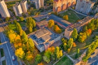 Aerial view of a residential area with several multi-story apartment buildings, surrounded by green spaces and trees with autumn foliage. The buildings have light-colored exteriors, and the area also includes roads, sidewalks, and recreational areas.