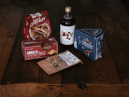 A collection of Swedish food items is arranged on a rustic wooden table. It includes a bag of gifflar pastries, a box of Annas Original Ginger Thins, a bottle of Skånska Spritfabriken cherry cordial, a box of Leksands Crispbread, and a small packet of snack mix.