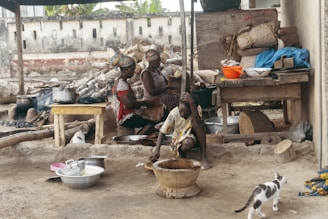 Three people are engaged in food preparation in an outdoor kitchen setting. They are surrounded by various cooking utensils, pots, and wooden tools. A cat is walking in the foreground near bowls and a wooden mortar, while the background shows a rustic wall and storage area with items like firewood and baskets.