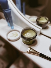 A tub of green cream labeled 'ARTISTRY' is open and placed on a bathroom counter near a mirror. Next to it, there is a brush with a white textured applicator head and a handle that has some text. There are also two blue tube containers in the background. The lid of the cream tub is placed beside it.