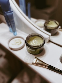 A tub of green cream labeled 'ARTISTRY' is open and placed on a bathroom counter near a mirror. Next to it, there is a brush with a white textured applicator head and a handle that has some text. There are also two blue tube containers in the background. The lid of the cream tub is placed beside it.